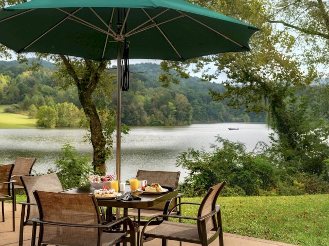 Outdoor dining setup with a table, chairs, and umbrella overlooking a lake and forested hills. Plates of food and drinks are on the table.