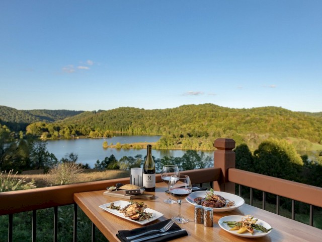 A scenic outdoor dining table with plates of food and wine overlooks a lush, green landscape and a lake under a clear blue sky.