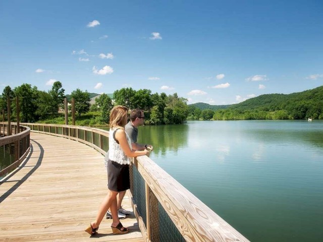A couple stands on a wooden boardwalk over a lake, surrounded by lush green hills and a clear blue sky.