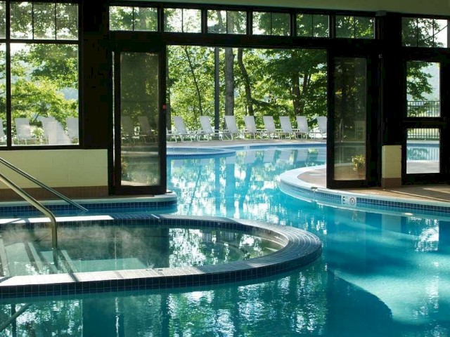 An indoor pool area with large windows, a hot tub, and a view of the outdoor pool surrounded by trees and lounge chairs.