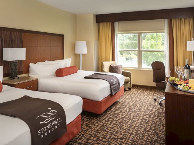 The image shows a hotel room with two beds, chairs, a window, and a table with wine and fruit. The room is labeled 