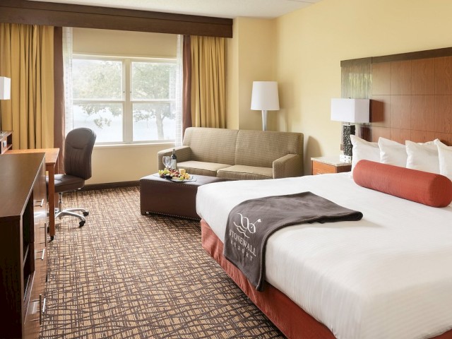 A modern hotel room with a large bed, sofa, desk chair, and TV, featuring warm decor and a window with a view.