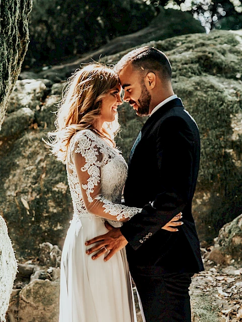 A couple in wedding attire embraces in a natural outdoor setting with rocks, moss, and greenery around them.