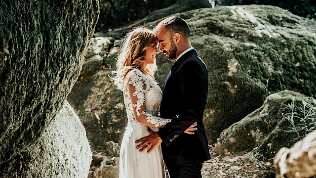 A couple in wedding attire embraces in a natural outdoor setting with rocks, moss, and greenery around them.