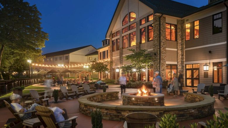 An outdoor patio of a large house with people sitting around a fire pit at night, under string lights, with chairs and trees nearby.