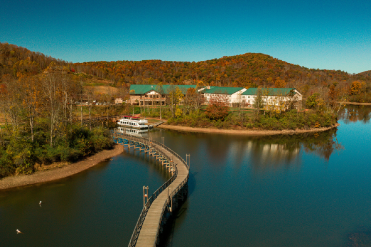 A scenic view of a lake with a curved boardwalk leading to a building surrounded by autumn trees and hills under a clear blue sky.