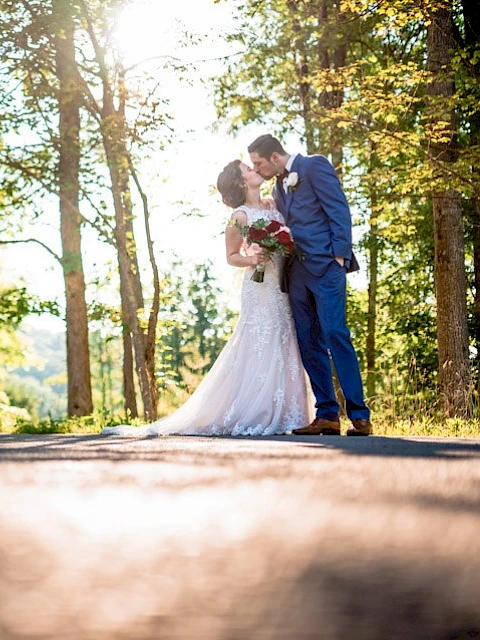 A newlywed couple shares a kiss in a sunlit forest, dressed in wedding attire, celebrating their special day outdoors.