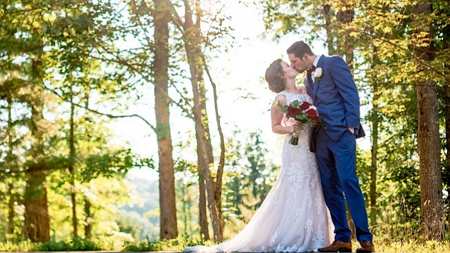 A newlywed couple shares a kiss in a sunlit forest, dressed in wedding attire, celebrating their special day outdoors.