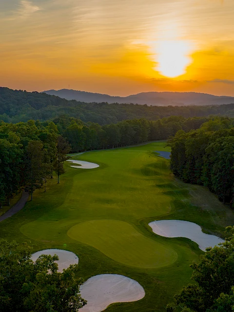 A beautiful aerial view of a golf course at sunset, surrounded by lush trees and rolling hills.