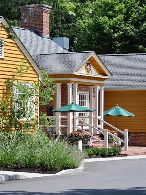 A charming yellow house with white trim, featuring a brick chimney, surrounded by lush green trees and a well-maintained garden.