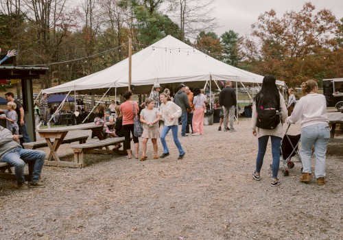 People are enjoying a lively outdoor market under a large white tent with vendor stalls, surrounded by fall foliage and picnic tables.