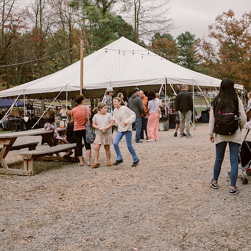 People are enjoying a lively outdoor market under a large white tent with vendor stalls, surrounded by fall foliage and picnic tables.