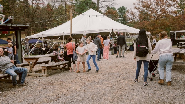 People are enjoying a lively outdoor market under a large white tent with vendor stalls, surrounded by fall foliage and picnic tables.