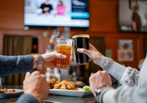 People are toasting drinks in a bar while enjoying food, with the background showing TVs and a cozy atmosphere.