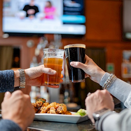 People are toasting drinks in a bar while enjoying food, with the background showing TVs and a cozy atmosphere.