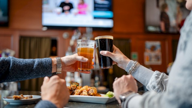 People are toasting drinks in a bar while enjoying food, with the background showing TVs and a cozy atmosphere.
