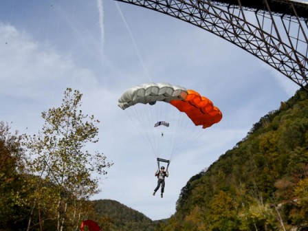 A person is skydiving under a parachute near a bridge and a wooded hillside on a cloudy day.