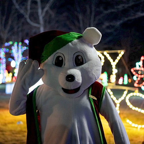 A mascot dressed as a dog in Christmas attire waving in front of colorful holiday lights at night.