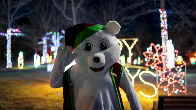 A mascot dressed as a dog in Christmas attire waving in front of colorful holiday lights at night.