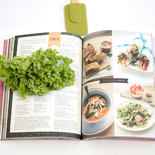 A cookbook open to lunch recipes, with fresh greens and vegetables surrounding it on a white surface.