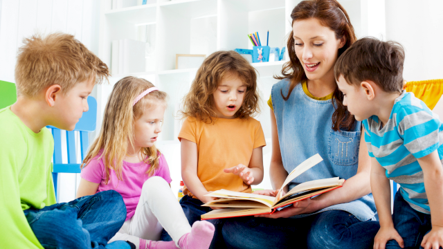 A teacher reads a storybook aloud to five children sitting on the floor in a bright, colorful classroom.