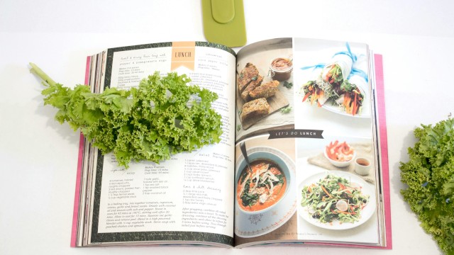 A cookbook open to lunch recipes, with fresh greens and vegetables surrounding it on a white surface.